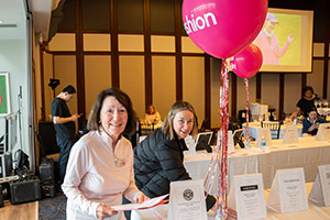 Two women smile standing at a large table with two pink baloons