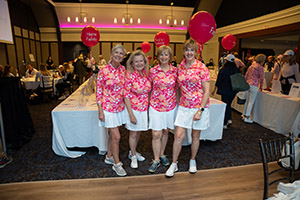 Four women wearing matching white skirts and pink shirts stand smiling