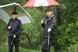 Two golfers wearing black stand holding umbrellas and golf clubs
