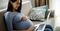 Pregnant woman sits on couch holding her stomach. She is looking at a laptop.