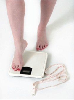 An image of a woman stepping onto a scale, with a tape measure at her feet. She must be having a ton of fun!