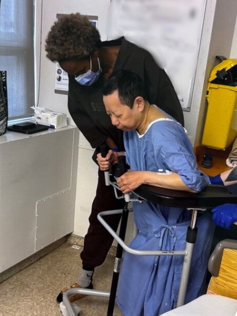 A woman with short black hair wearing a blue hospital gown, holds onto a walker with the help of a woman wearing all black who is supporting her upright