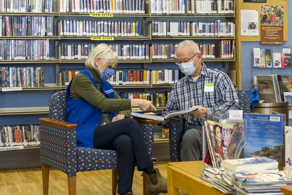 A patient and volunteer both in face masks interacting with a book between them in front of a library.