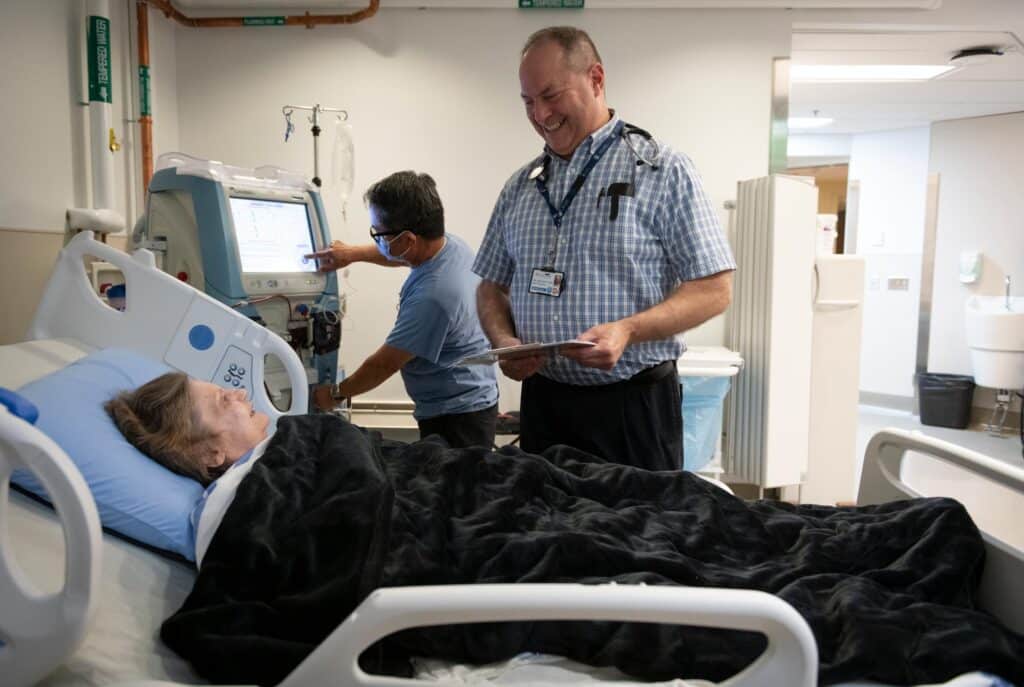 Doctor Tobe stands talking to a patient laying in a hospital bed. A technician is seen in the background looking at a monitor with the patients vitals